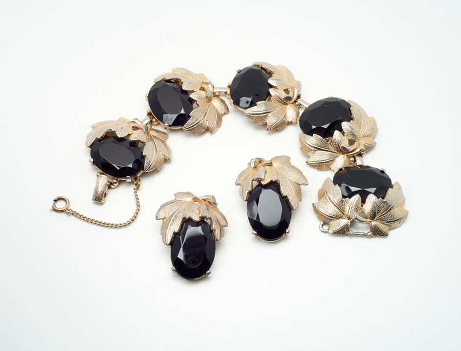 Elsa Schiaparelli, Bracciale e Orecchini, 1950 ca.Bracciale e orecchini a clip, omaggio a Bacco, con pietre nere sfaccettate montate su foglie di vite in metallo dorato che accompagnano grappoli d’uva realizzati con cristallo noir. Metallo placcato oro, pietre di vetro